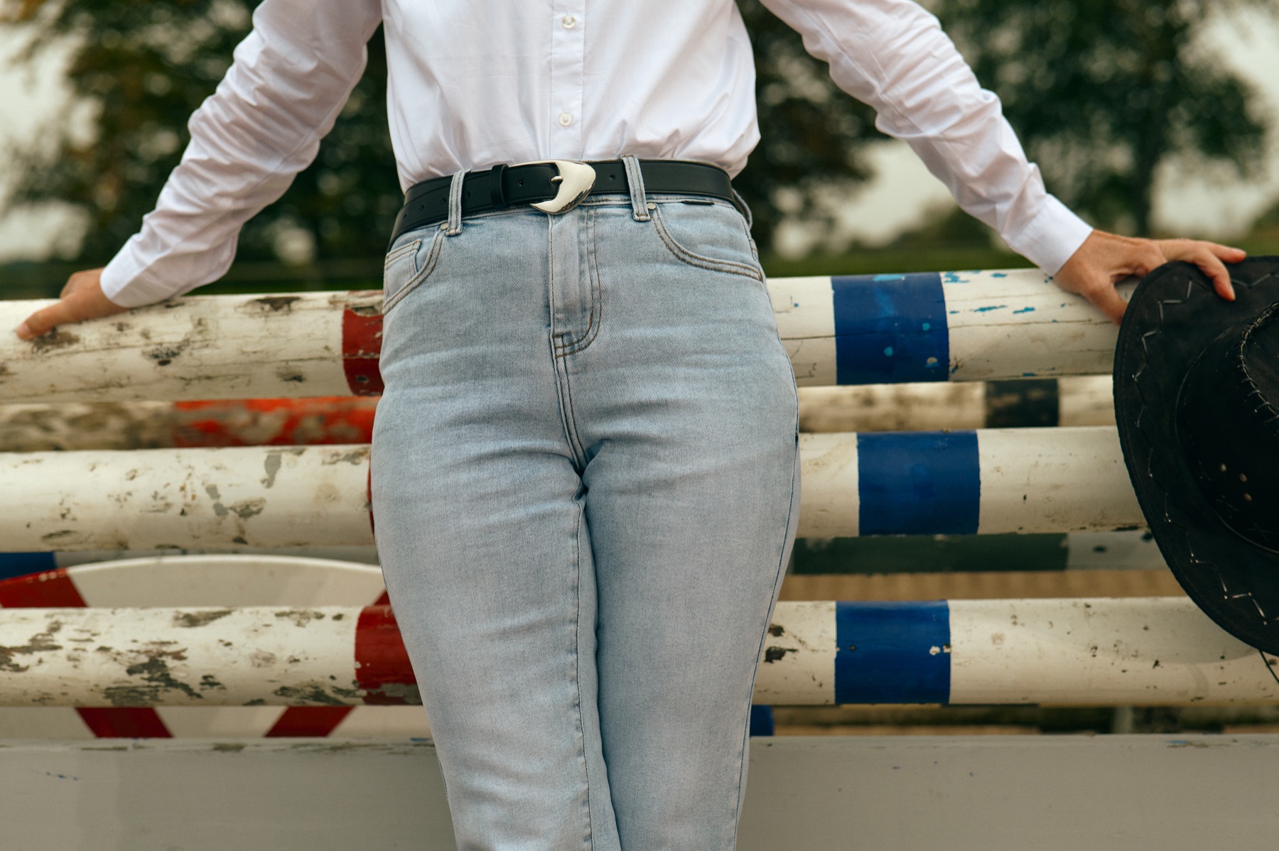 Person in white shirt and light jeans leaning on a striped fence, holding a black hat.